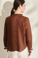 Blusa de lino color marrón con escote en V, cuello con volados y mangas largas con puños fruncidos que presentan detalles en color crudo, al igual que en los hombros. Cuenta con cierre frontal de botones.