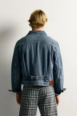 Campera de denim de algodón en color azul, con lavado efecto desgastado, cuello solapa y cierre frontal de botonadura metálica. Presenta bolsillos con solapa en el pecho, mangas largas con puños abotonados y trabilla ajustable en la cintura posterior.