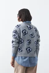 Cardigan de tejido jacquard color marrón con estampado floral en tonos rojo y rosado. Presenta cuello redondo, cierre frontal con botones y terminaciones acanaladas en puños y ruedo.