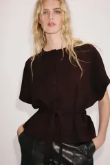 Chaqueta marrón de punto con cuello redondo y manga corta. Presenta cierre frontal con botones y un cinturón integrado del mismo tejido con lazo para atar en la cintura.