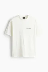 Remera de algodón de peso medio, color blanco hueso, con cuello redondo y hombros caídos. Presenta un pequeño estampado en cursiva negra en el pecho.