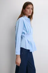 Camisa blanca de popelín con cuello solapa y manga larga, cintura entallada con pliegues y cierre frontal con botones ocultos bajo solapa.