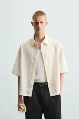 Camisa de corte relajado (relaxed fit) color crema o blanco roto, confeccionada en tejido con mezcla de algodón. Presenta un bordado sutil en relieve con patrón de ondas o líneas diagonales entrecruzadas. Tiene cuello solapa, manga corta y un bolsillo de plastrón en el pecho.