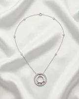 Collar de acero con cadena fina que presenta pequeños apliques de circonias a lo largo de su extensión. El dije central tiene un diseño circular doble, con el borde exterior pavé de circonias y tres pequeñas piedras rojas incrustadas en la parte inferior del círculo interior.
