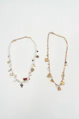 Set de dos collares con dijes variados. El primero cuenta con una cadena de perlas blancas y dijes metálicos que incluyen un cactus, cerezas rojas, un corazón y una luna. El segundo collar presenta una cadena dorada con cuentas pequeñas y dijes metálicos con formas de corazón, flores y elementos geométricos.