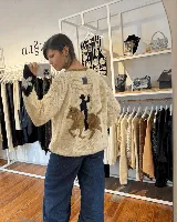 Buzo de punto tejido a mano en lana merino, color beige claro con diseño en la espalda que representa a un gaucho montando a caballo bajo una nube. Presenta mangas largas con detalles en contraste.