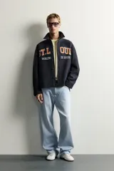 Sobrecamisa de ajuste relajado en color azul marino, con cuello de solapa, manga larga con puño abotonado y cierre frontal con botones. Presenta bordados contrastantes en el frente con la inscripción "St. Louis Worldwide Goods" en tonos naranja y blanco.
