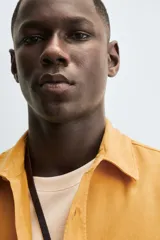 Camisa de hombre color mostaza, con cuello solapa, manga larga y dos bolsillos plastrón con solapa en el pecho. Presenta un calce relajado y cierre frontal de botonadura.