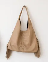 Cartera estilo hobo de cuero sintético color beige, con flecos laterales, detalle de tachas metálicas verticales en el frente y aplique con estrellas grabadas en la base.