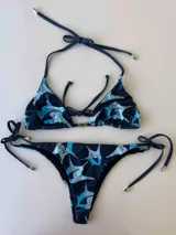 Bikini de dos piezas con corpiño triangular fruncido y bombacha regulable. Presenta una estampa abstracta en tonos azul oscuro y celeste, con detalles metálicos en las terminaciones de las tiras.