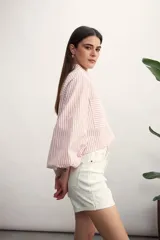 Camisa a rayas verticales color beige y blanco, con cuello clásico, cierre frontal con botón dorado, mangas largas abullonadas con puños elásticos, dos bolsillos frontales y corte holgado.