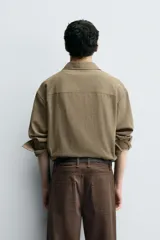 Camisa de manga larga color marrón verdoso, con cuello solapa y cierre frontal de botonadura. Presenta un tejido con textura rústica y un calce relajado.