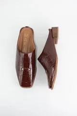 Zueco de punta almendrada confeccionado en cuero con textura croco. Presenta un diseño de talón abierto, taco cuadrado bajo y detalle de dos tachas metálicas en la parte superior.