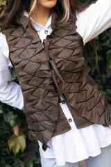 Chaleco de tweed marrón con textura, presenta un diseño frontal con volados estructurados y cordones decorativos en zigzag. Cuenta con cierre frontal mediante lazos ajustables.