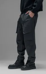 Pantalón de estilo utilitario confeccionado en textil técnico ripstop color gris oscuro. Presenta un corte oversize con cintura elástica, cierre frontal con broche técnico, bolsillos delanteros de gran tamaño con cierre y sistema de ajuste en los tobillos mediante broches snap.