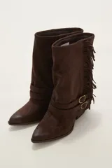 Bota corta de gamuza color chocolate, con punta fina y taco bajo. Presenta detalle de flecos en el lateral exterior y dos correas decorativas con hebillas metálicas en el tobillo.