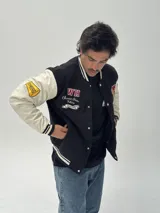 Campera estilo varsity con cuerpo negro y mangas blancas. Presenta parches bordados en el pecho y las mangas, cierre frontal con botones a presión y puños, cuello y ruedo acanalados con rayas blancas y negras.