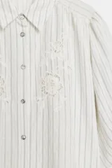 Camisa blanca a rayas sutiles, de cuello solapa y manga abullonada por debajo del codo. Presenta delicados bordados florales perforados en el frente y cierre frontal con botones.