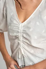 Blusa marrón de algodón con bordados sutiles, escote en V, mangas cortas abullonadas y detalle de fruncido con cordón ajustable en el centro del frente.