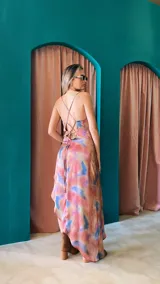 Vestido largo de crepé con estampado floral en tonos rosados, morados y blancos. Presenta escote buche con drapeado y espalda baja ajustable con tiras finas. El ruedo es irregular, más corto en el frente.