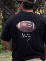 Remera de algodón negra con estampado de un balón de fútbol americano y la frase "Together, we are America". Incluye una firma en blanco debajo del balón.