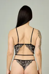 Conjunto de lencería compuesto por un bralette triangular y una tanga, confeccionados en encaje negro con detalles florales en color hueso. El bralette presenta breteles ajustables y diseño con tiras decorativas, mientras que la tanga cuenta con doble tira lateral regulable.