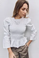 Blusa blanca de algodón con cuerpo fruncido y elástico, cuello redondo con detalle de puntilla bordada, mangas largas abullonadas con puños elásticos y ruedo con volado.
