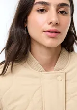 Campera acolchada de color beige con diseño matelasseado, cuello, puños y dobladillo en rib elástico. Cuenta con bolsillos frontales con solapa y cierre de botones a presión.