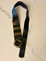 Faja de cuero negro con diseño envolvente, hebilla metálica cuadrada en tono bronce envejecido y detalles de varillas metálicas horizontales decorativas.
