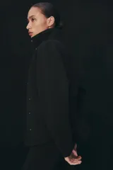 Chaqueta negra de manga larga con cuello subido, bolsillos delanteros y cierre frontal con botones ocultos bajo solapa.