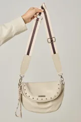 Bandolera de cuero vacuno color beige, con diseño de solapa curva decorada con tachas metálicas. Incluye una correa regulable de cinta con franjas oscuras y cierre principal con tirador largo.