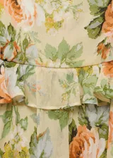 Vestido corto de gasa color amarillo pálido con estampado de rosas anaranjadas y hojas verdes. Tiene escote recto con volantes, tirantes finos y cierre lateral con cremallera.