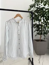 Camisa de lino blanca con rayas verticales negras, cuello mao y cierre frontal con botones marrones.