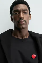 Camiseta de manga larga color negro, corte relajado (relaxed fit) con cuello redondo y un pequeño parche bordado en el pecho con un dado rojo y corazones blancos.