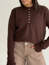Sweater de punto color marrón, con cuello redondo y cierre frontal parcial con botones. Presenta mangas largas con puños terminados en volados.