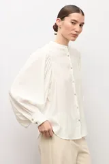 Camisa blanca de corte recto con cuello mao, cierre frontal de botones y mangas largas abullonadas con puños ajustados.