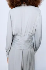 Camisa de escote en pico con lazada frontal en el mismo tejido, manga larga con puños y bajo drapeado. Presenta un acabado satinado y cierre lateral con cremallera oculta.