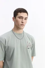 Remera de manga corta color verde salvia, con cuello redondo y un estampado gráfico en la espalda que incluye tipografía en rojo y blanco, y fotografías en blanco y negro.