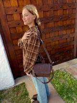 Cartera bandolera de cuero color chocolate con solapa frontal, costuras a la vista y herrajes metálicos. Incluye dos correas desmontables, una corta y una larga, y cuenta con bolsillo interior.