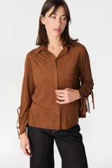 Camisa de color camel con textura geométrica en relieve, cuello clásico y cierre frontal con botones. Presenta mangas largas con detalle de tiras ajustables y aberturas en los puños.
