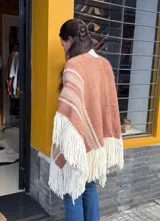 Poncho tejido en telar de peine en lana merino de color terracota y blanco, con diseño geométrico de rayas y grandes flecos en los bordes inferiores.