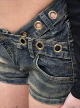Short de denim tiro bajo con diseño cruzado en la cintura, efecto desgastado y detalles de ojalillos metálicos dorados.