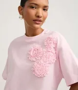 Remera blanca de algodón con cuello redondo y corte boxy, con apliques de flores de tejido en relieve en la zona del pecho.