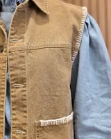Chaleco de denim color marrón, confeccionado en 100% algodón, con bolsillos frontales amplios, cierre de botones y costuras decorativas bordadas en blanco en los bordes y los bolsillos.
