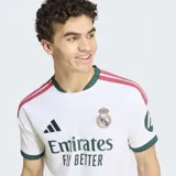 Camiseta de futbol Adidas del Real Madrid, primera equipacion. Diseño en color blanco con detalles en verde oscuro en el cuello y los puños, y franjas fucsias en los hombros. Cuenta con el logo de la marca, el escudo del club bordado y el patrocinador oficial en el frente.