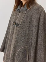 Capa de tejido de punto en color gris melange, con diseño abierto y cierre frontal mediante un botón tipo duffle de madera con detalle en cuero. Presenta bolsillos laterales amplios y bordes con costuras a la vista.