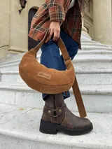 Cartera tipo hobo de cuero gamuzado color camel, con correa ajustable y etiqueta de cuero con logo grabado en el frente.