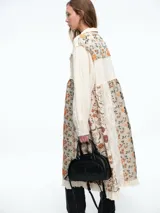 Chaqueta larga de diseño patchwork en tonos grises, con combinación de texturas que incluyen paneles de tela lisa, tela texturizada blanca y paneles con estampado floral en relieve. Presenta cierre frontal con botones, cuello camisero y bordes deshilachados.