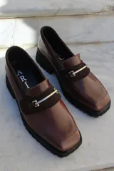Mocasín de cuero vacuno color bordó con suela track negra de goma. Presenta un diseño de punta cuadrada con un herraje metálico plateado sobre una banda de gamuza en la capellada.