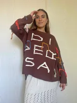 Sweater de punto color marrón con diseño oversize, cuello redondo y mangas largas. Presenta bordados artesanales en relieve con hilos de colores y detalles en lurex, incluyendo flecos decorativos distribuidos por toda la prenda.